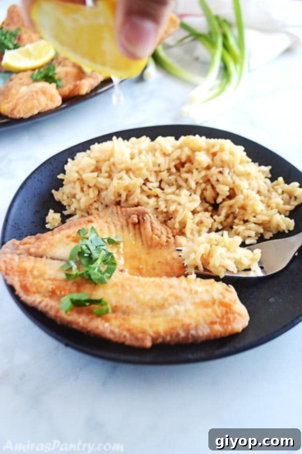 A hand gently squeezing a fresh lemon wedge over a piece of pan-fried tilapia, presented on a black plate with brown rice and a fork.
