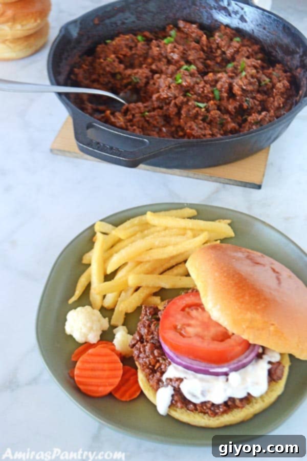A sloppy joes sandwich with white shawarma sauce , tomato and onion rings on a agreen plate with some fries on the side.