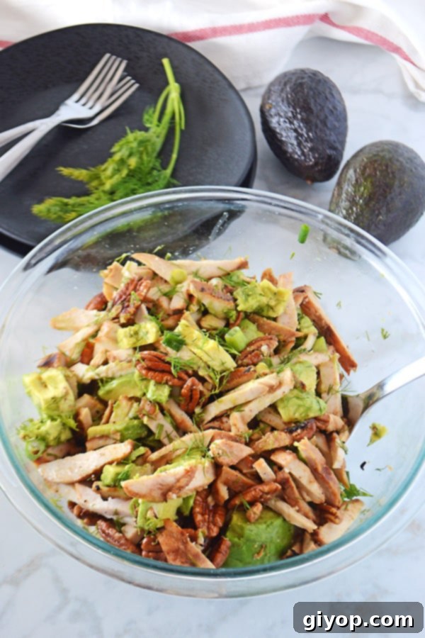 A large, vibrant serving bowl overflowing with avocado chicken salad, surrounded by black plates and forks, with two whole, ripe avocados strategically placed as a garnish, ready for a feast.