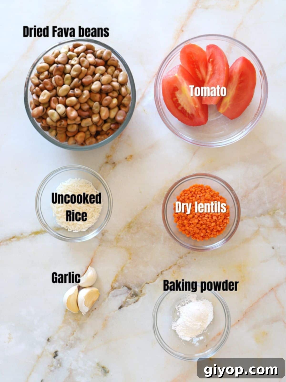 Various ingredients for Ful Medames laid out on a marble countertop: dry fava beans, rice, red lentils, a Roma tomato, and garlic cloves.