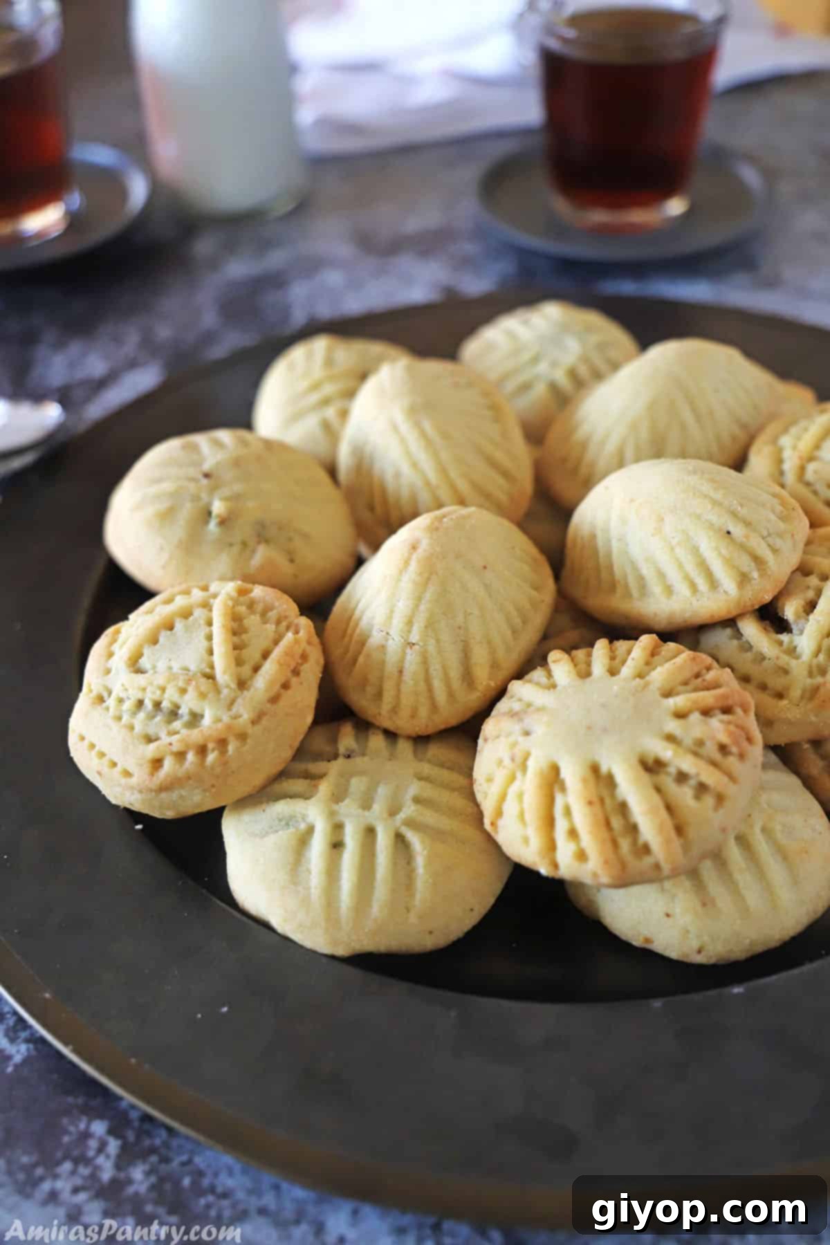 mamoul cookies on a black metal plate with some cups of tea and milk in the back.