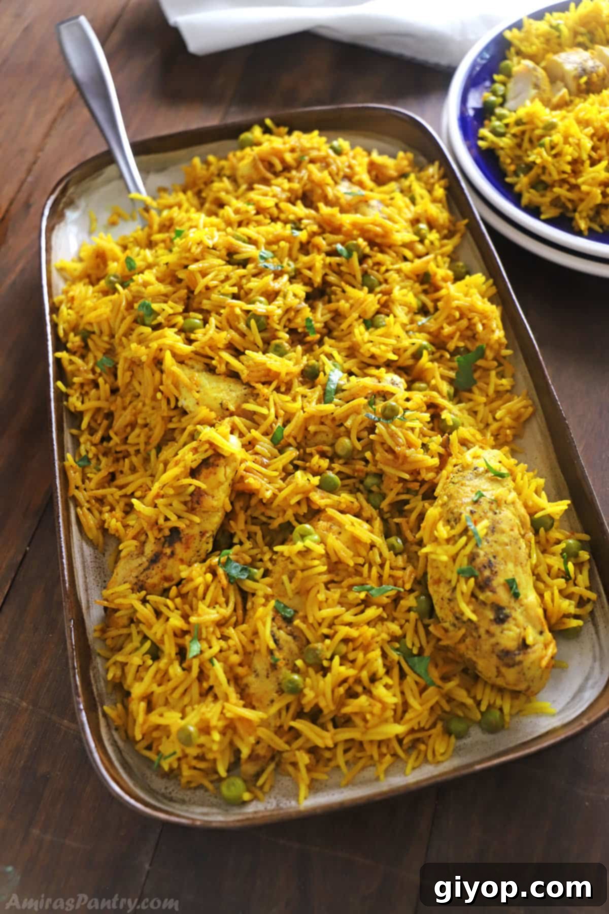A large serving platter of golden yellow rice with succulent chicken pieces and green peas, garnished with fresh herbs, placed on a rustic wooden table, ready to be served.