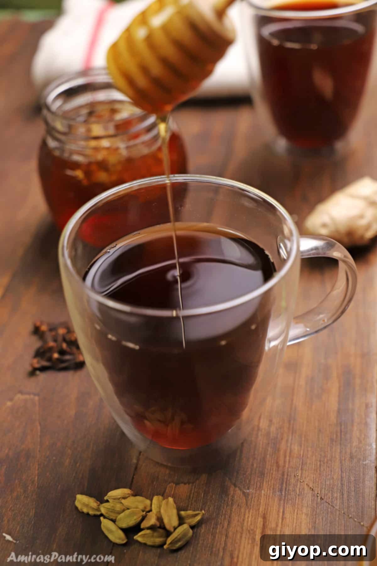 A honey spoon pouring honey into a cup of cardamom tea, with steam rising from the cup.