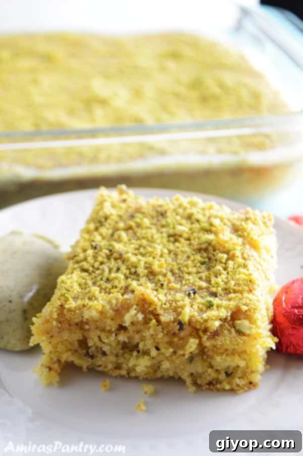 A slice of vibrant green pistachio semolina cake on a white plate, garnished with more pistachios and a delicate white flower. The background shows hints of a soft, romantic setting.