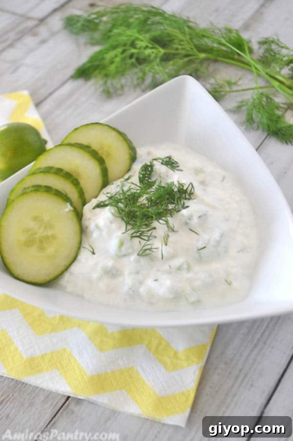 Tzatziki sauce served on a white triangular plate with sliced cucumber on the side. A lemon and dill weed are on the back of the photo.