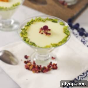 Four elegant glass cups of creamy Middle Eastern milk pudding, Mahalabia, garnished with green pistachios and red rose petals, arranged on a dark wooden tray.