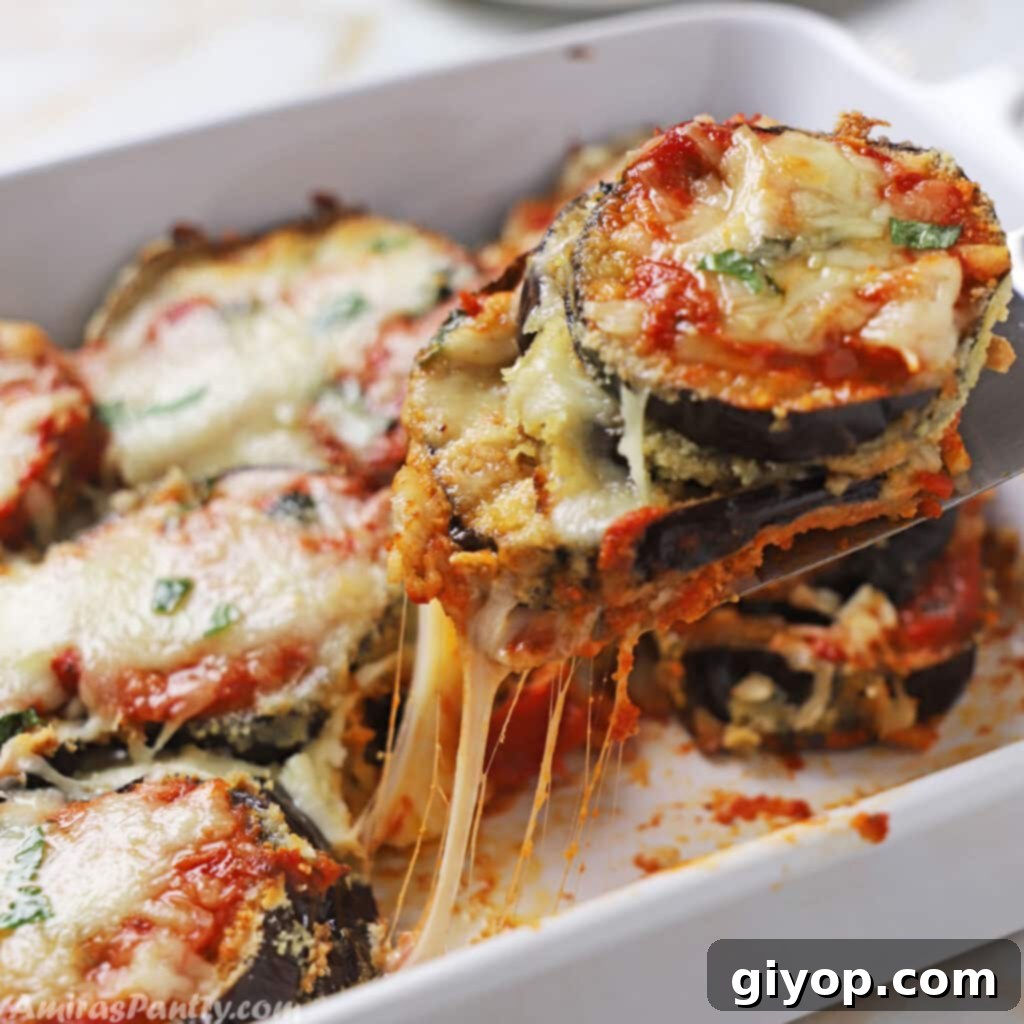 A hand expertly scooping a generous portion of golden-brown eggplant parmesan from a white deep casserole dish, revealing layers of cheese and sauce.