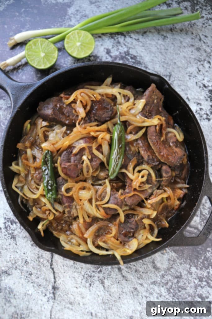 Sizzling beef liver and caramelized onions in a cast-iron skillet, garnished with fresh lemon wedges and chopped green onions.