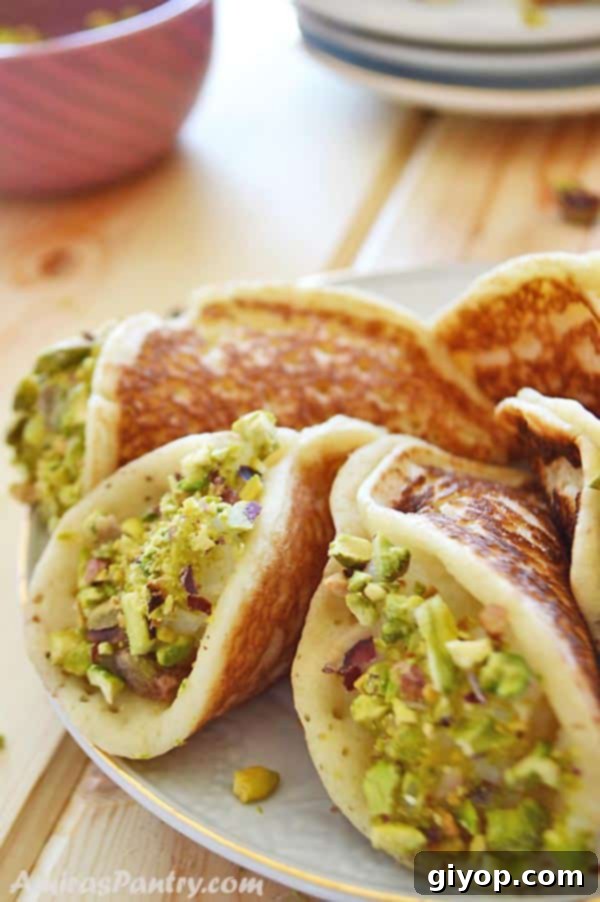 A white plate with several stuffed Qatayef Asafiri, dipped in crushed pistachios, with a small bowl of extra pistachios in the background.