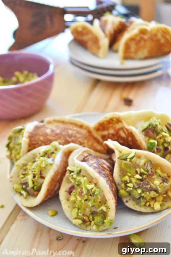 A white plate filled with Qatayef Asafiri stuffed with cream, accompanied by a small bowl of crushed pistachios in the background.