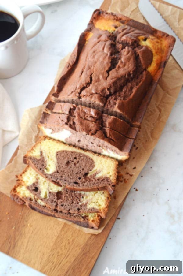 A loaf of marble cake on a wooden cutting board with third of it being cut on the board with a coffee mug on the side. This easy chocolate marble loaf cake is great for beginners.