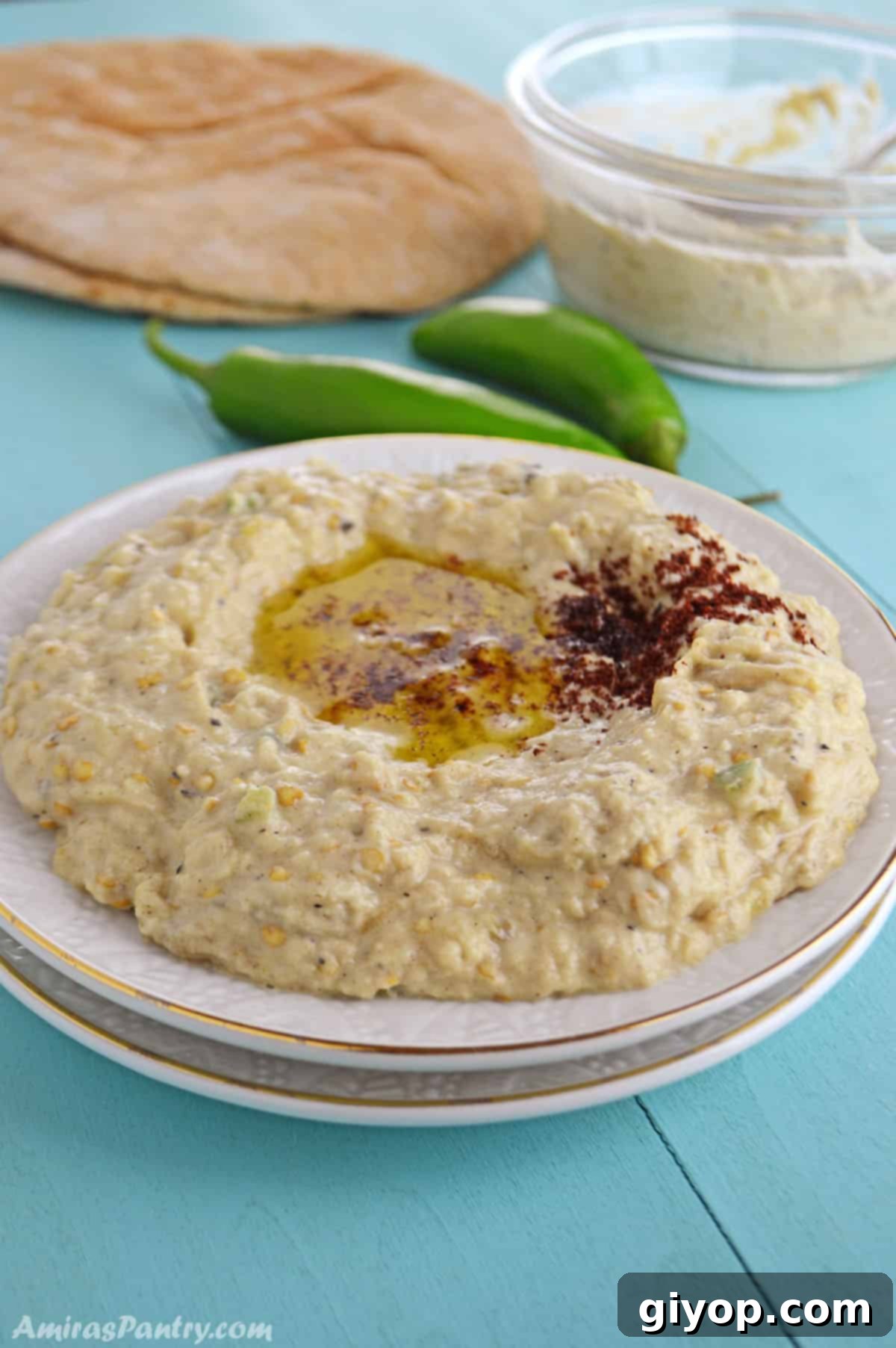 Baba Ghanoush elegantly presented on a small white serving plate, garnished with olive oil and parsley, alongside fresh pita bread on a light blue wooden tabletop.