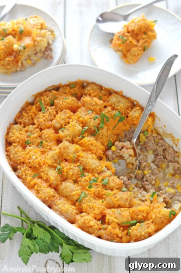 Tater tot in a white oven dish with a serving spoon in it and two servings of the dish on small white plates on the side on a wooden table.