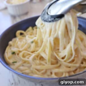 Kitchen tongs scooping some pasta with alfredo sauce from a skillet.