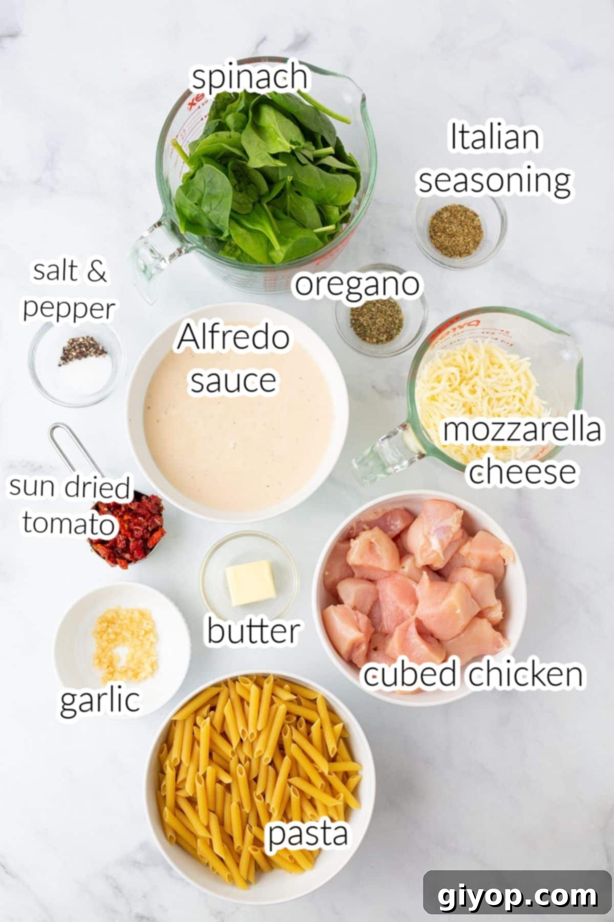 Various ingredients for a creamy pasta dish, including pasta, chicken, spinach, sun-dried tomatoes, and cheese, neatly arranged on a marble countertop.