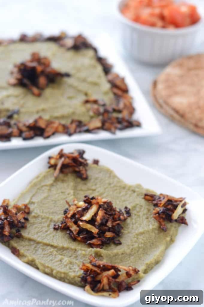 Two white plates of Egyptian Bissara, a creamy fava bean dip, garnished with crispy fried onions, served alongside a small bowl of fresh tomato and warm pita bread.