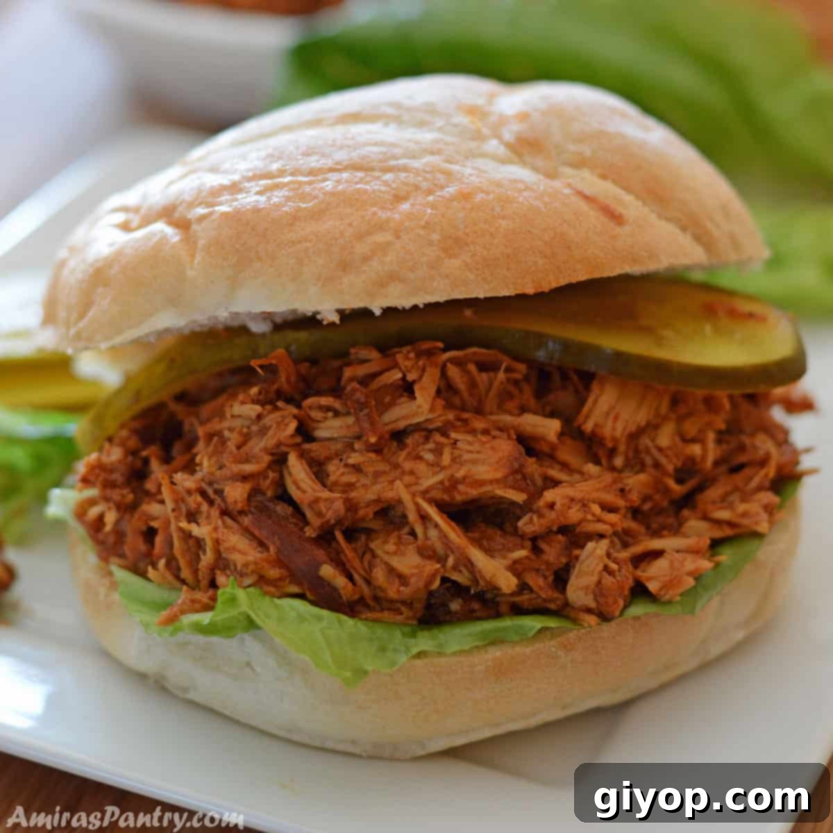 A mouthwatering pulled chicken sandwich on a white plate, showcasing layers of tender chicken and fresh garnishes.