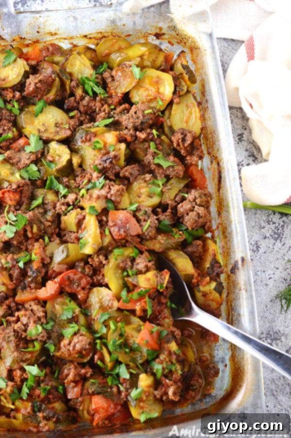 A pyrex glass baking dish filled with a golden-brown ground beef zucchini casserole, with a serving spoon.