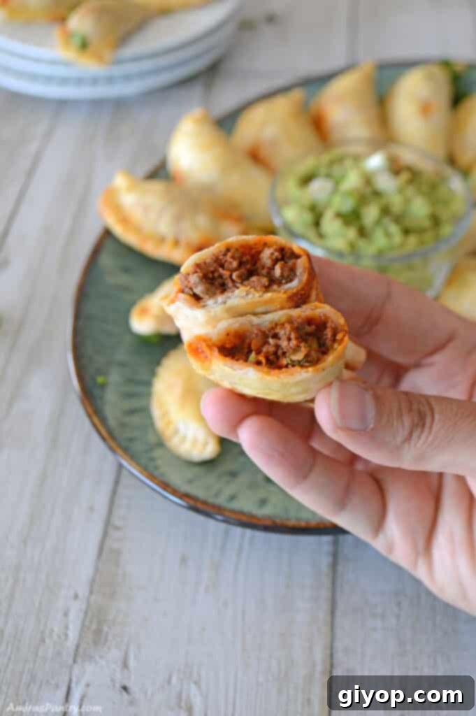A hand gently holding a golden-brown beef empanada, cut in half to reveal its savory filling.