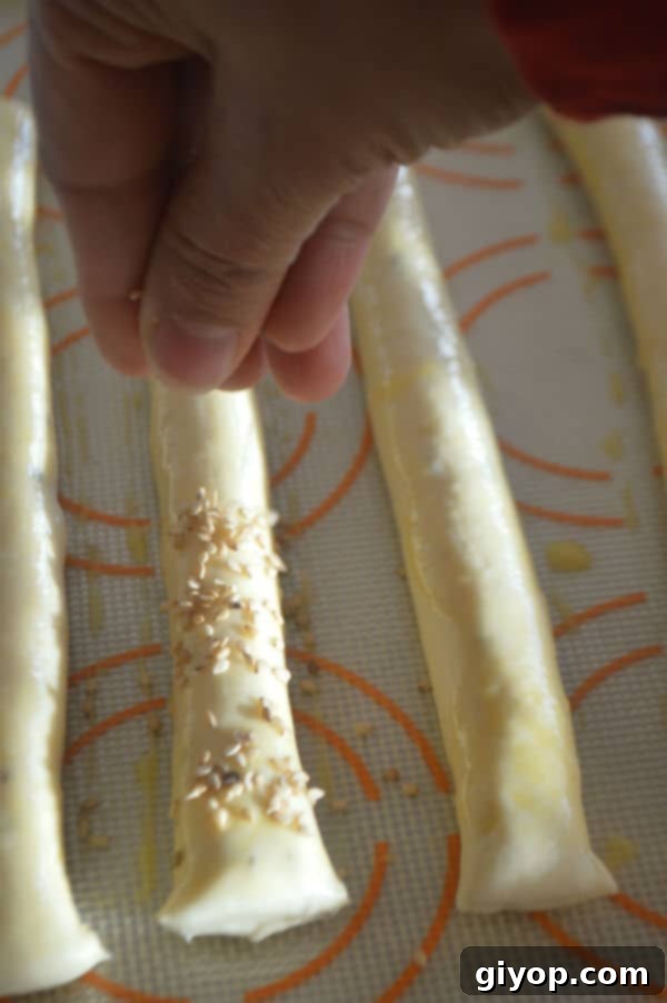 Crispy Sesame Breadsticks 8 A hand sprinkling sesame seeds over breadsticks arranged on a baking sheet, preparing for the oven.