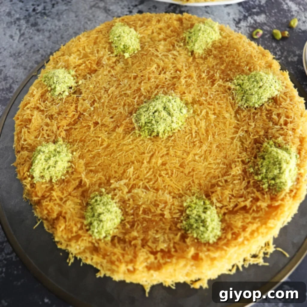 Knafeh garnished with crushed pistachios on a black metal platter, highlighting its golden crust and creamy interior.