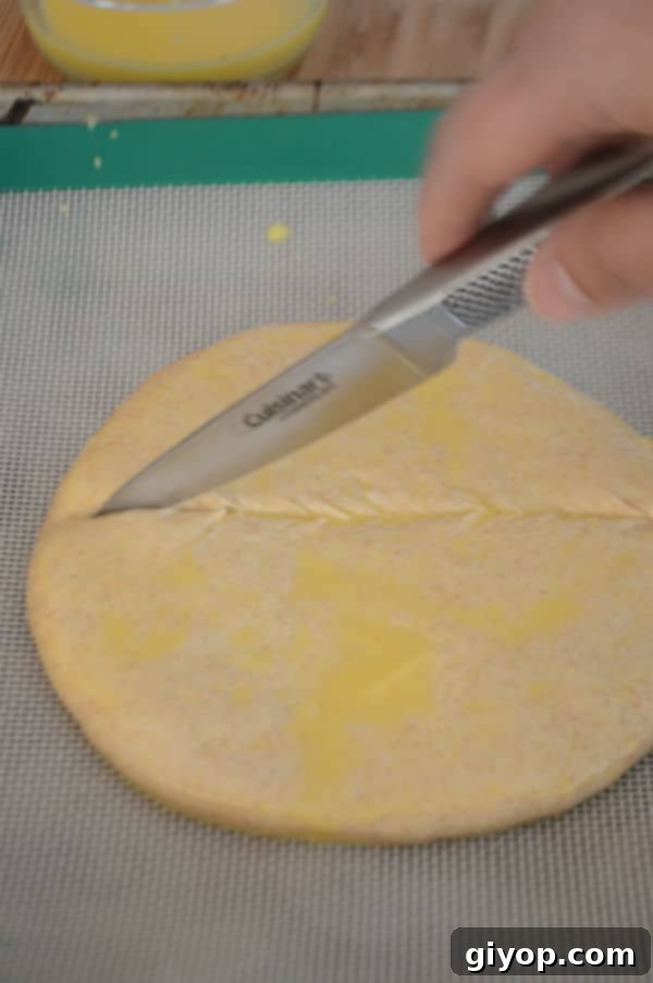 A close-up of a round dough disc on parchment paper, being scored with a sharp knife, and sprinkled with sesame seeds.