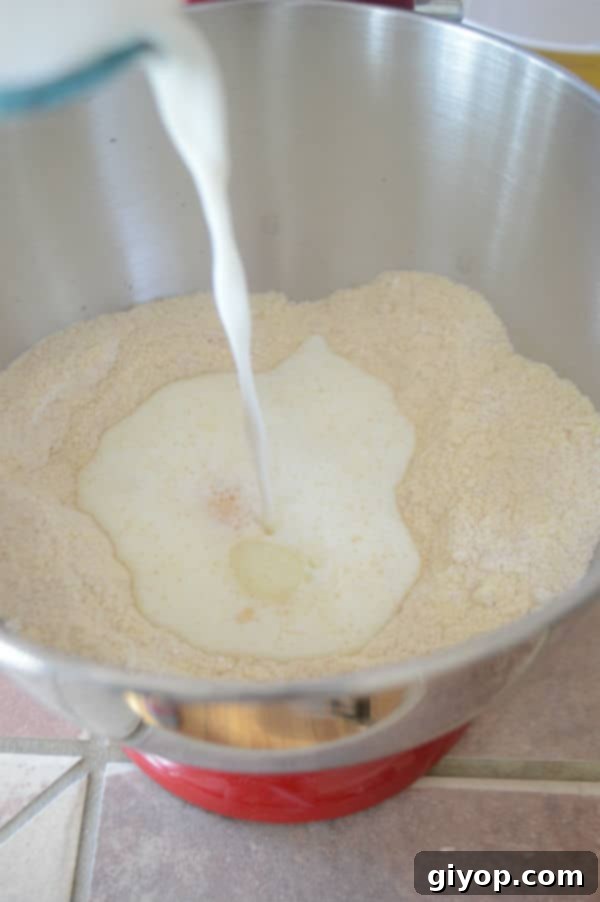 A metal mixing bowl with dough ingredients, as warm milk is poured in to begin forming the dough.