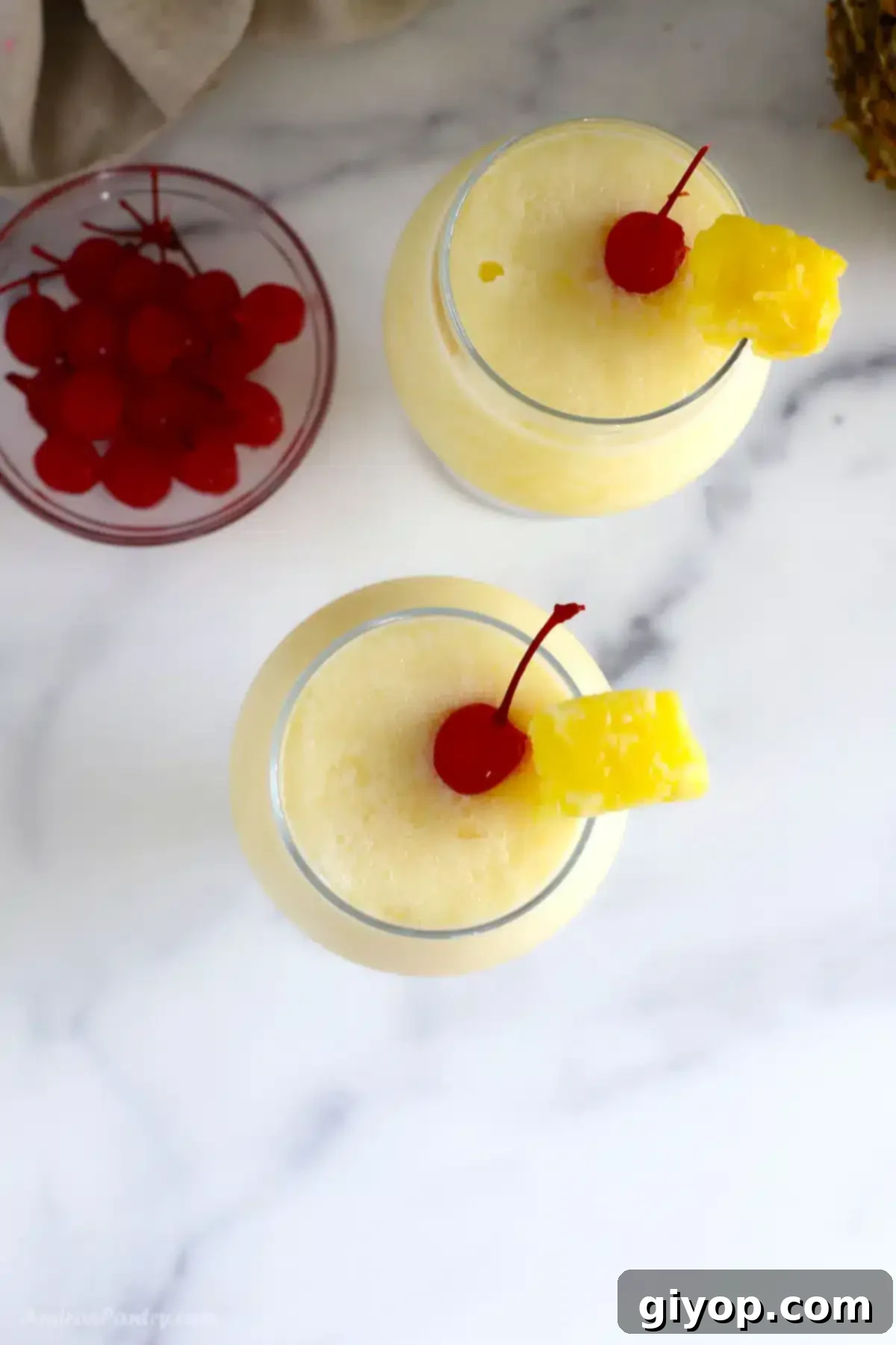 An overhead image of two glasses of Virgin Pina Colada, garnished with cherries, on a light surface.