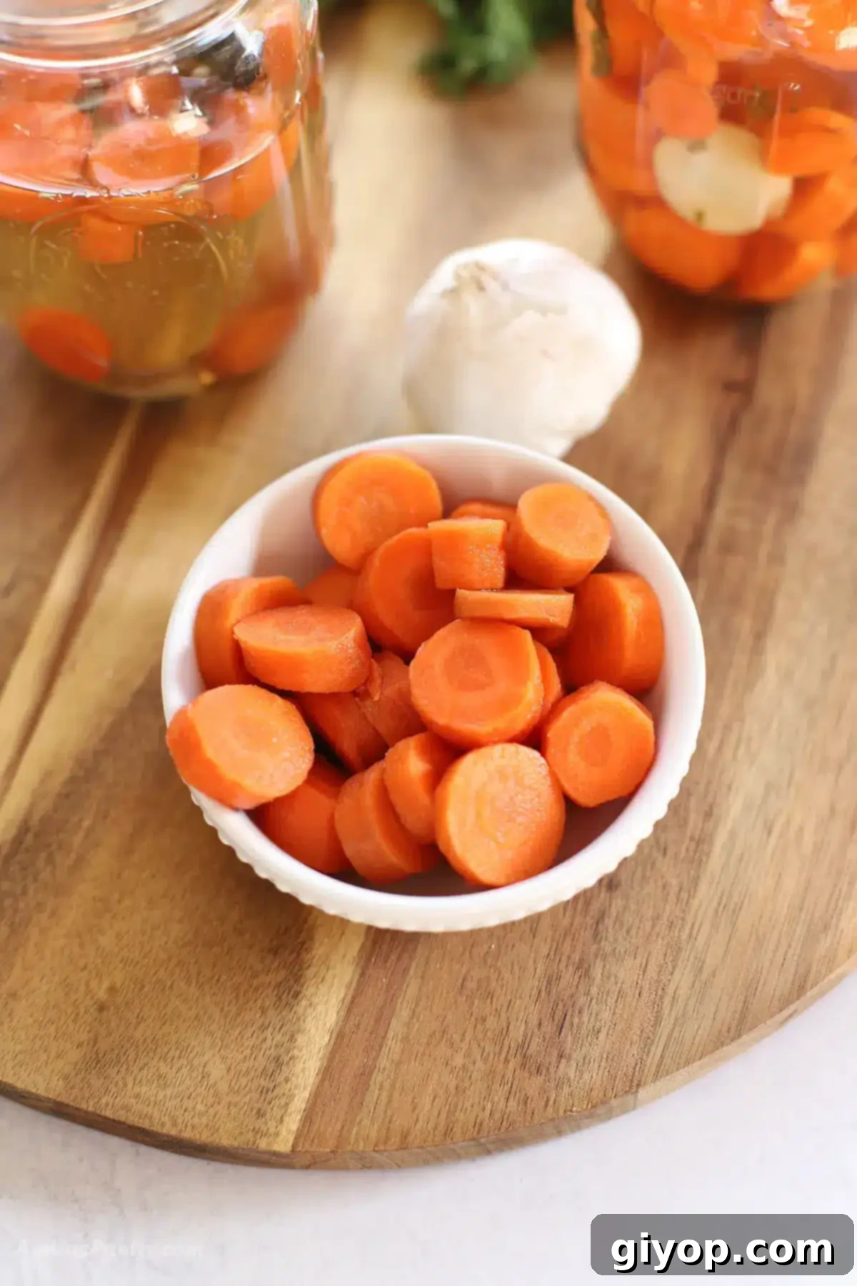 A serving of quick pickled carrots in a white bowl, accompanied by garlic cloves and fresh herbs in the background, highlighting their readiness to be enjoyed.