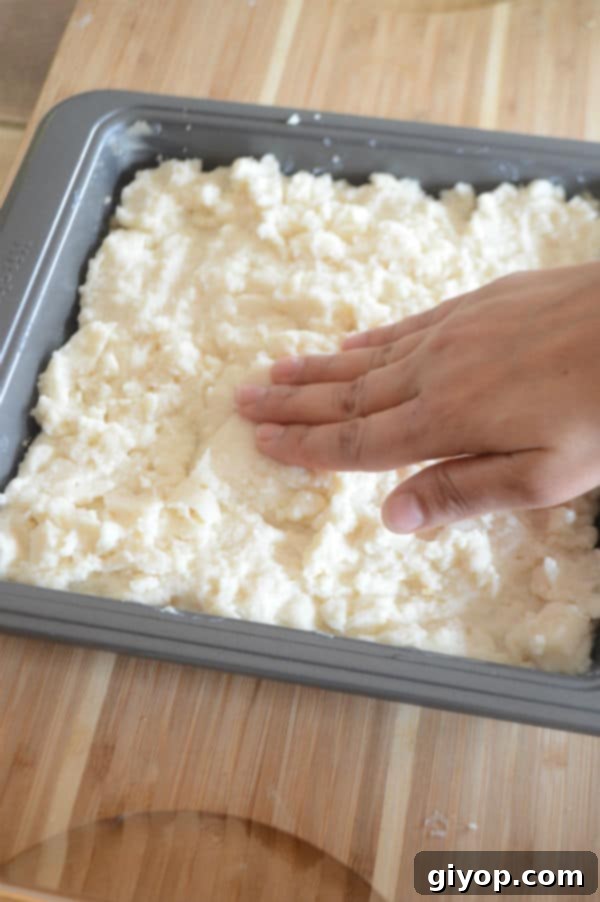 Pistachio-Packed Maamoul with Velvety Ashta 11 A hand spreading the creamy Ashta filling evenly over the semolina base in the baking dish, creating a smooth middle layer.