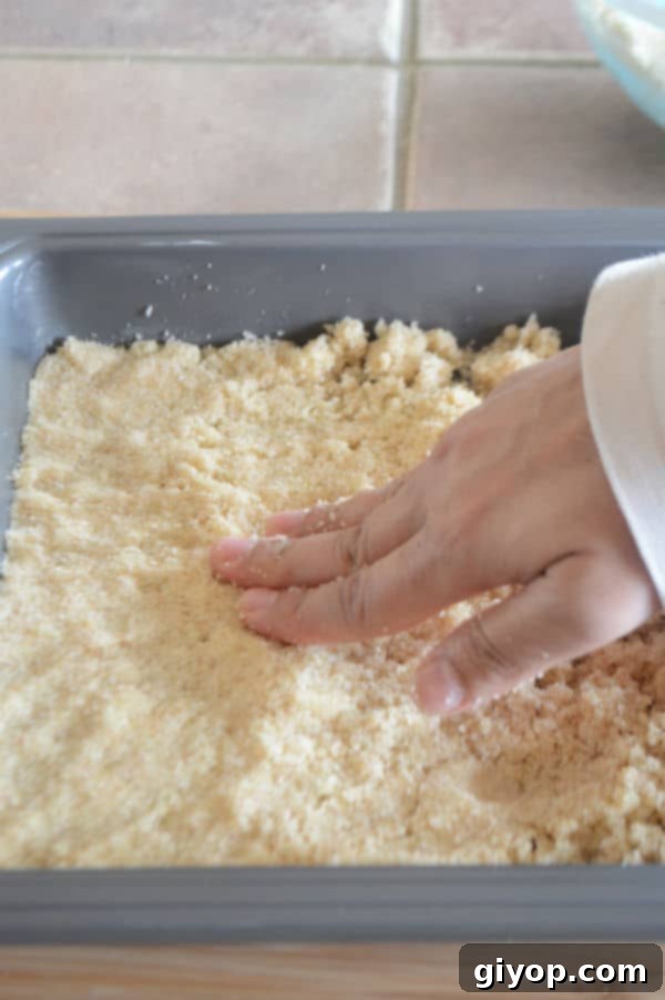 Pistachio-Packed Maamoul with Velvety Ashta 10 A hand pressing half of the prepared semolina dough mixture evenly into the bottom of a baking dish, forming the base layer.