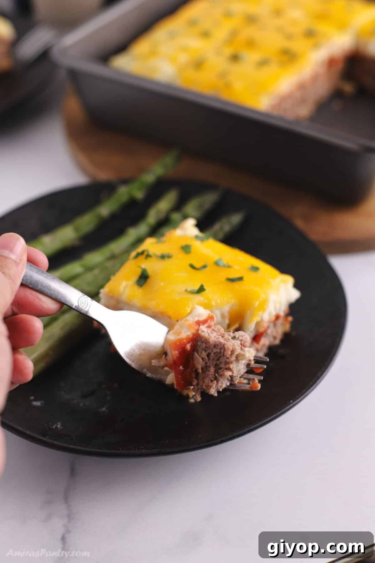 A fork cutting into a piece of Meatloaf Casserole, with the rest of the casserole in the background, showing its delicious layers.