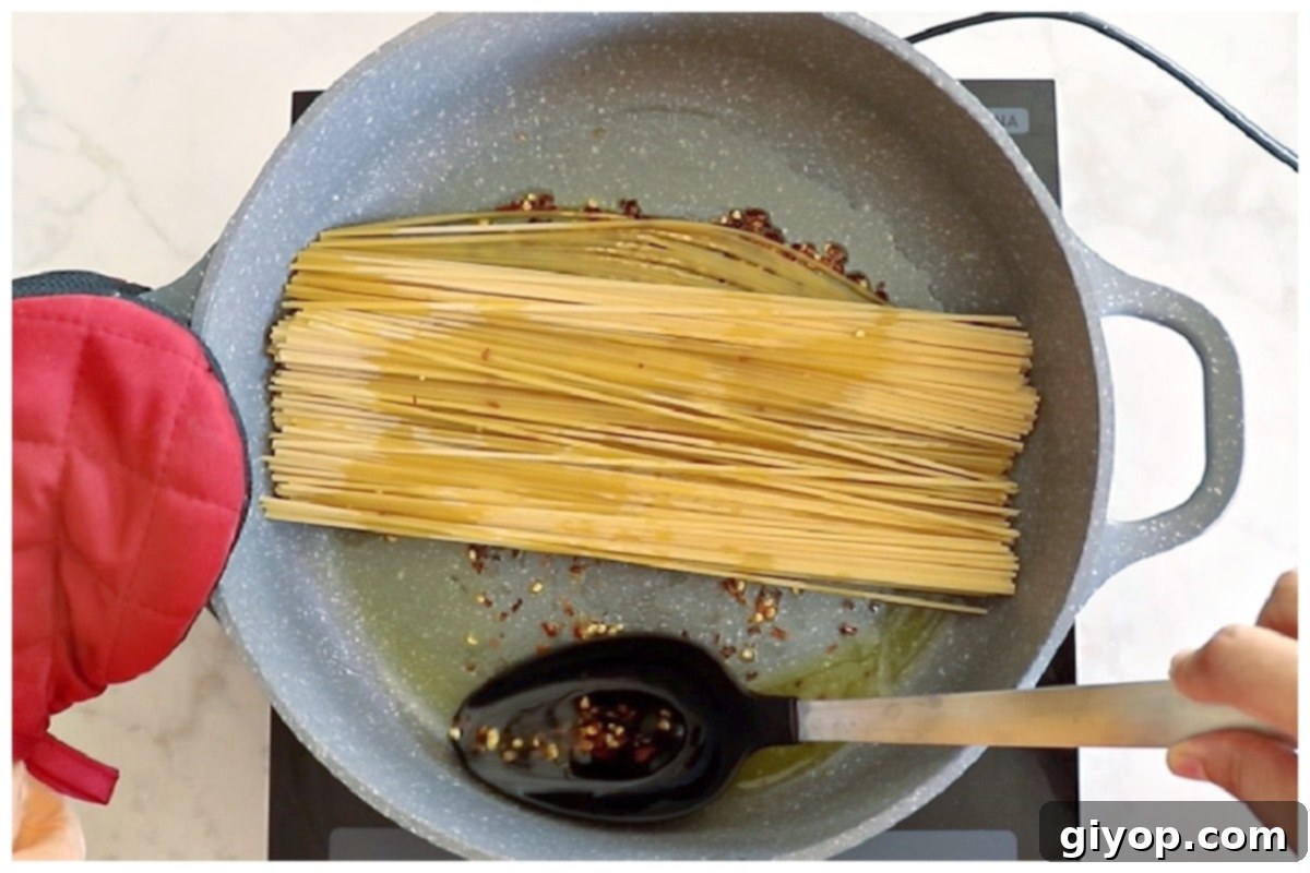 A chef using tongs to coat dry spaghetti in chili oil by tilting the pan.