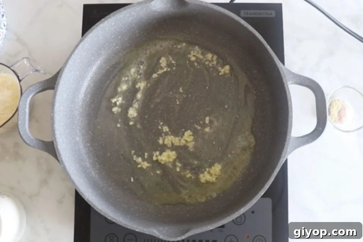 Minced garlic cloves sizzling gently in melted butter within a large skillet, creating a fragrant base for the Alfredo sauce.