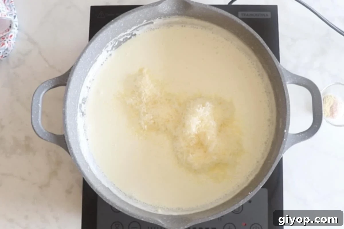 A large skillet showing the Alfredo sauce mixture being topped with a generous amount of freshly grated Parmesan cheese, ready to be stirred in.
