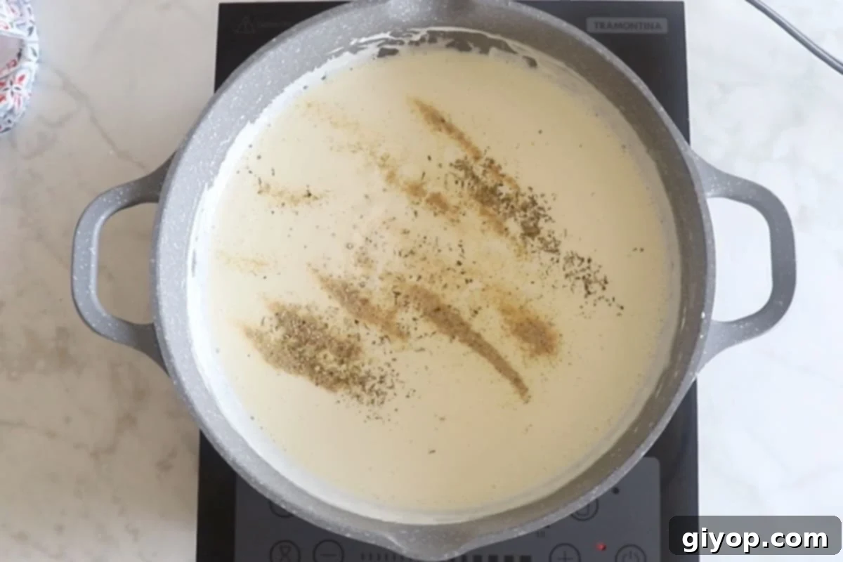 Seasonings being sprinkled into the simmering Alfredo sauce in a large skillet, adding depth of flavor before final thickening.