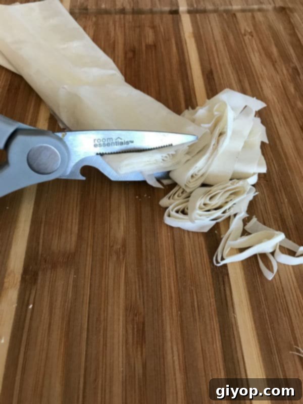 A scissor cutting strips of delicate phyllo pastry dough on a wooden table.
