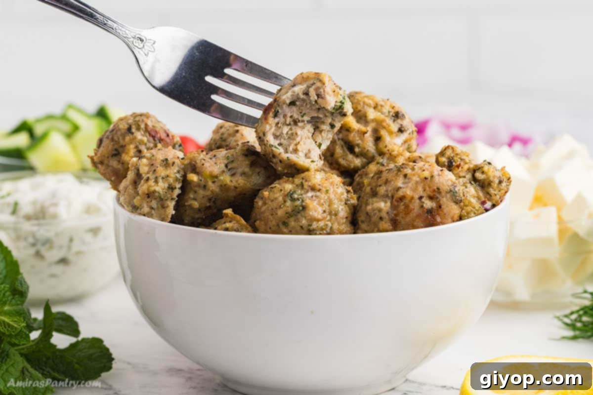 A fork holding one succulent Greek chicken meatball, freshly baked and still warm, with a small bite taken to reveal its juicy interior.