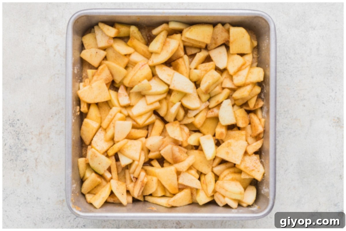 Decadent Salted Caramel Apple Pie Bars 11 A close-up view of thinly sliced, spiced apple filling beautifully arranged in an even layer over the baked shortbread crust in a square baking pan.