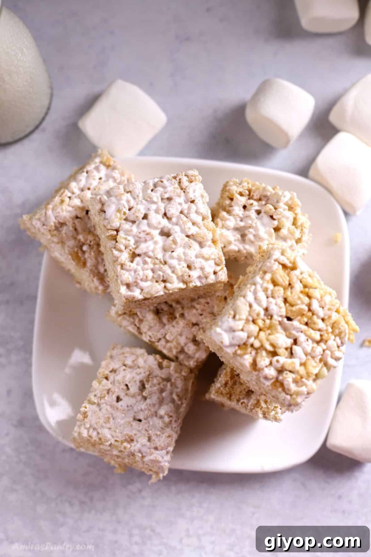 Delicious Rice Krispie squares on a pristine white plate, with soft marshmallows artfully blurred in the background, hinting at their gooey goodness.