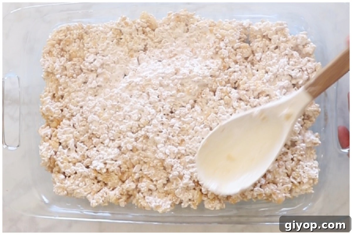 Freshly made Rice Krispie treats in a glass casserole dish, with a spatula gently pressing down to create an even, compact layer.