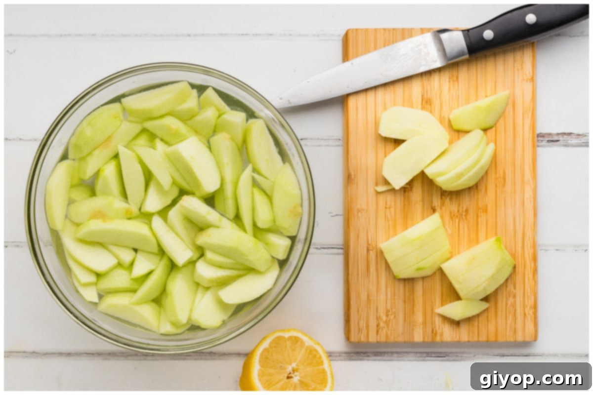 Neatly sliced apples soaking in a large bowl of lemon water, with some prepared apples on a wooden cutting board nearby.