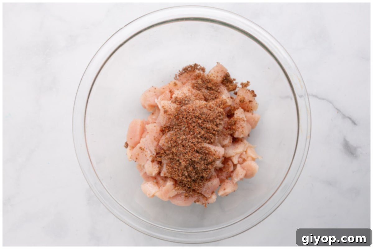 Cubed chicken tossed with a blend of spices in a clear glass bowl, showcasing even coating.