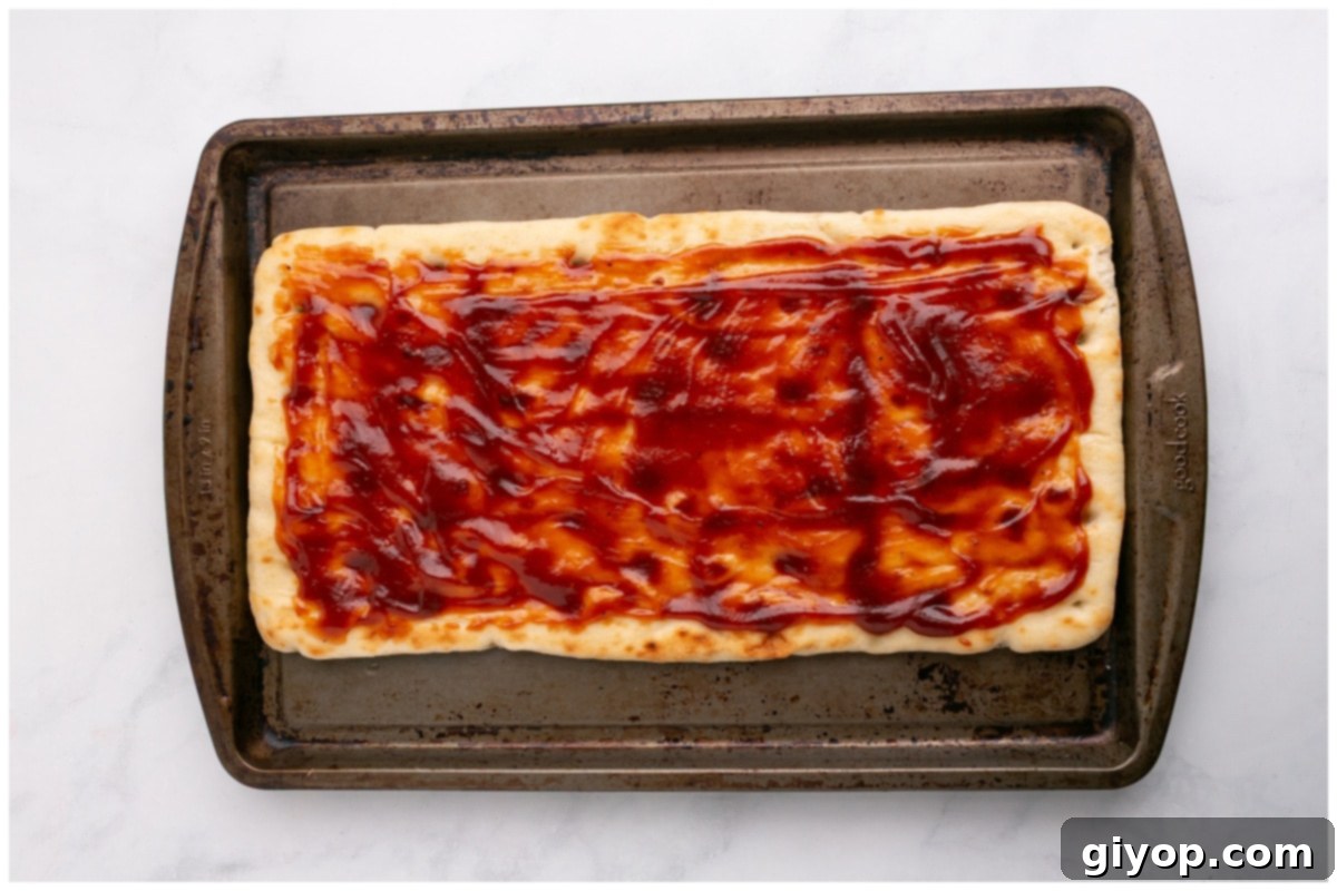 A baking tray holds flatbread bases that have been brushed with garlic oil and generously spread with BBQ sauce.
