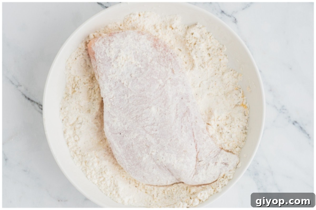 Hands carefully dredging a chicken breast in the seasoned flour mixture, ensuring an even coating.