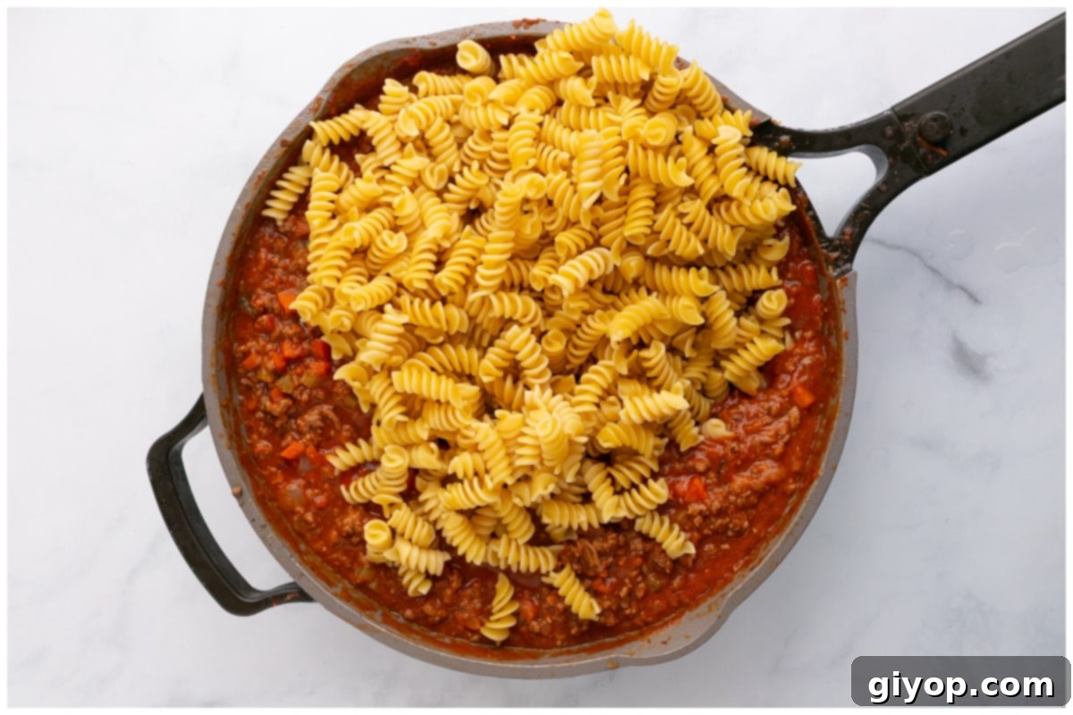 Cooked pasta being added to a large skillet containing the rich Bolognese meat sauce, ready to be tossed together for the baked dish.