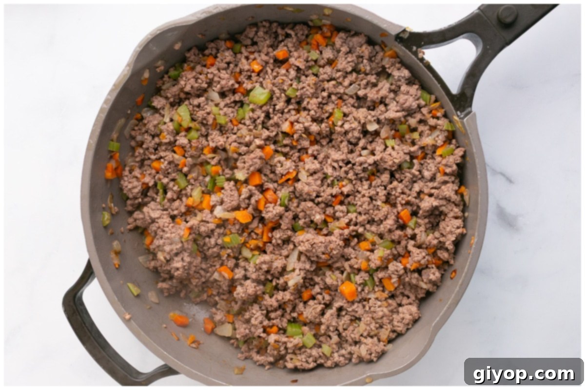 A large non-stick skillet containing a mixture of browned ground beef, along with the softened carrots, celery, and onions. The beef is finely crumbled and thoroughly cooked, preparing the base for the rich Bolognese sauce.