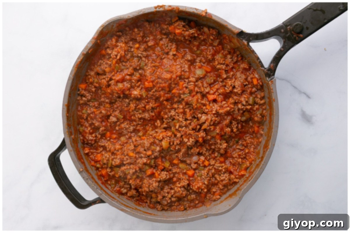 A large skillet filled with a rich meat sauce, including crushed tomatoes, beef broth, and herbs, simmering gently on the stove. The sauce is bubbling slowly, indicating it's developing deep flavors.