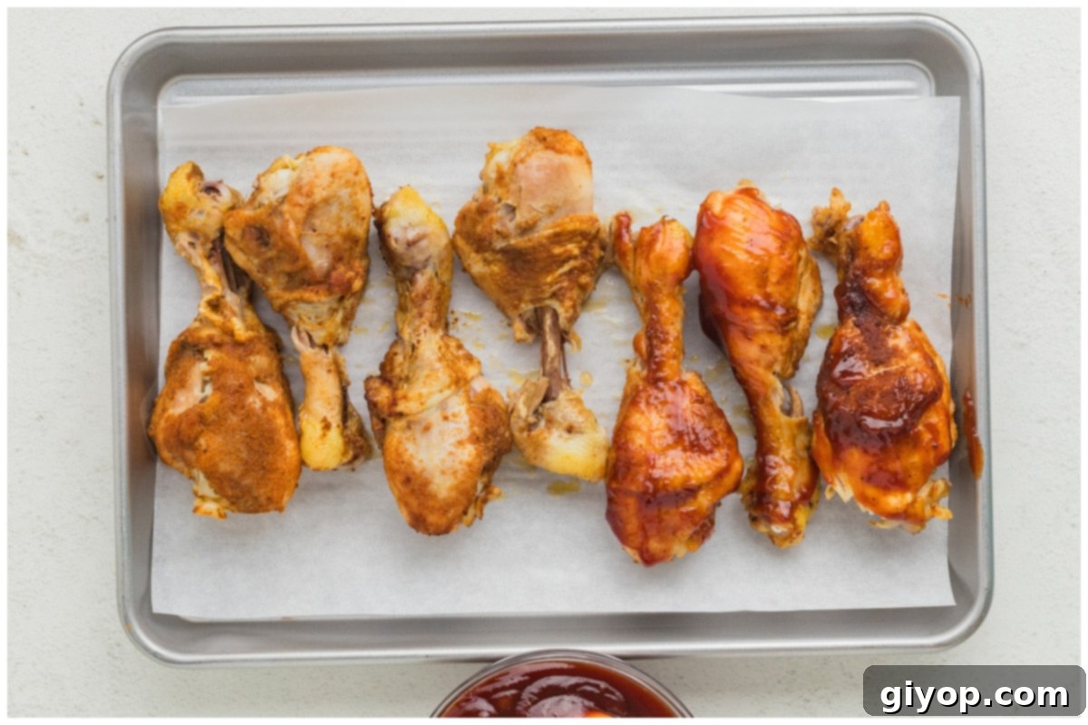 Cooked chicken drumsticks on a baking sheet, half brushed with glistening barbecue sauce, ready for broiling.
