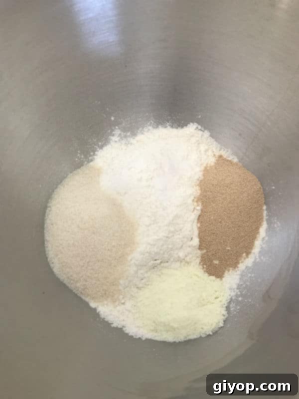 A metal mixer bowl with dry ingredients: flour, sugar, salt, yeast, and dry milk.